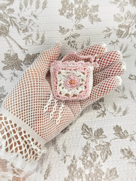 A small crocheted pink and white floral pouch or charm is held in a hand wearing a beige fishnet glove, set against a vintage floral fabric background.