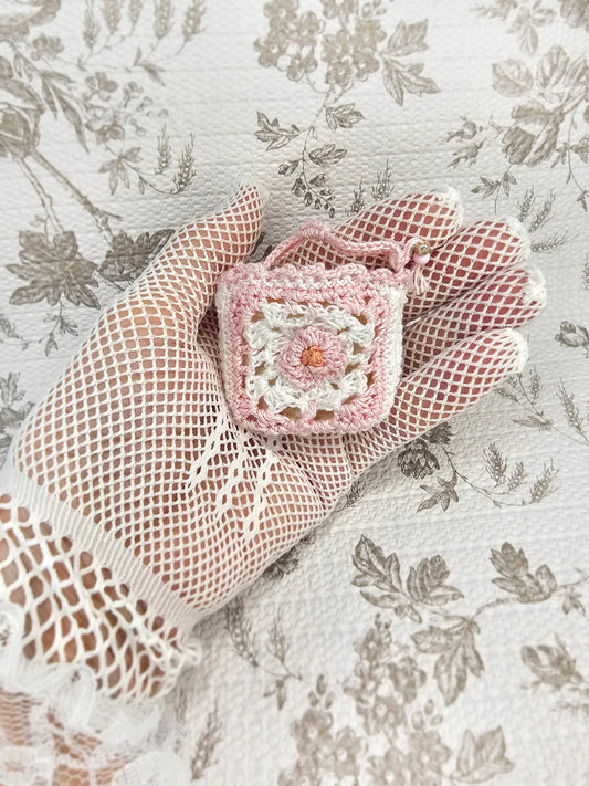 A small crocheted pink and white floral pouch or charm is held in a hand wearing a beige fishnet glove, set against a vintage floral fabric background.