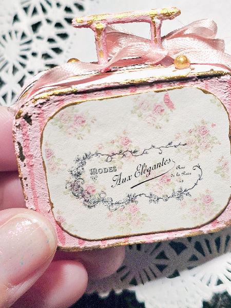 A miniature vintage-style pink suitcase ornament or decorative piece held by a hand, featuring a pink ribbon handle, gold trim, and a floral label that reads 'Mодель aux Elegantes'.