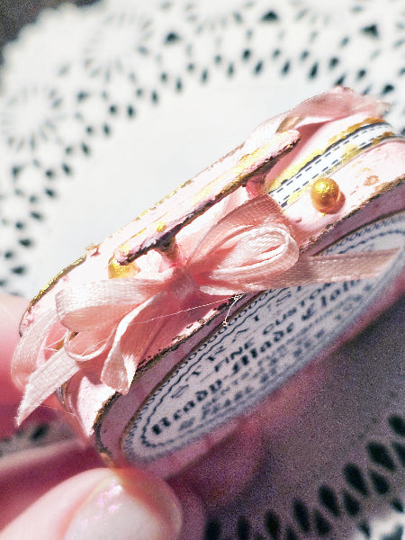 A close-up of a pink decorative ribbon-wrapped item with a bow, gold foil accents, and a printed oval label, held by a hand over a white lace doily.