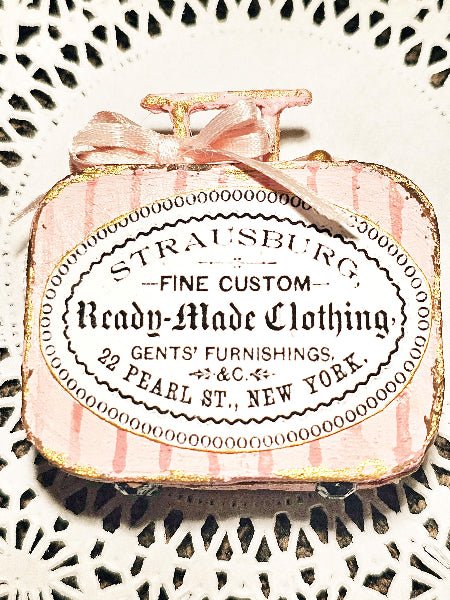 A vintage-style decorative miniature suitcase ornament or box with a pink and white striped exterior, gold glitter trim, a small gold handle, and a pink ribbon bow. The front features a large white oval label with black text reading 'Strausburg Fine Custom Ready-Made Clothing' and additional decorative text, shown on a white lace doily background.