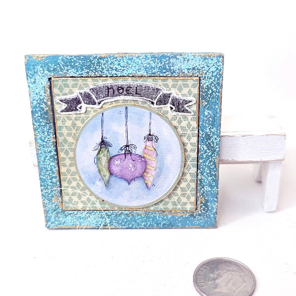 A small square decorative plaque or framed art piece with a distressed blue border and a central illustration of three hanging hearts in pastel colors under a banner reading “noel.” It is photographed on a white background with a small white stand and a coin for scale visible.