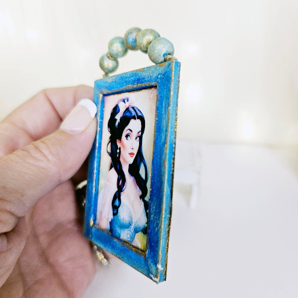 Hand holding a small framed artwork of a woman with long hair against a white background