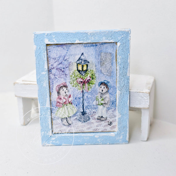 A small square framed art piece with a distressed light blue frame, featuring a whimsical winter scene of two children under a decorated streetlamp. The frame is displayed on a white surface with miniature white stools in the background.