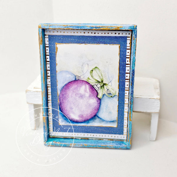A square framed art print featuring a watercolor illustration of a purple ornament and a white bow, set in a distressed blue frame and displayed on a small white stand against a white background.
