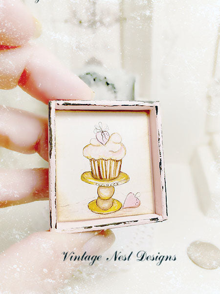Small framed artwork of a cupcake on a stand held by a hand with a blurred background.