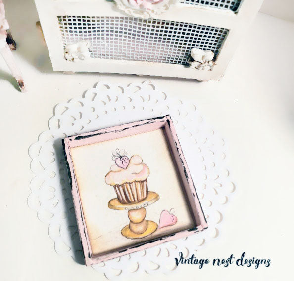 Framed illustration of a cupcake on a stand with hearts, placed on a decorative white surface.