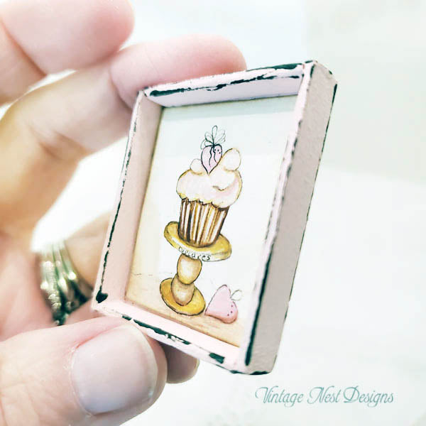 Small framed artwork of a cupcake held by a hand with a white background