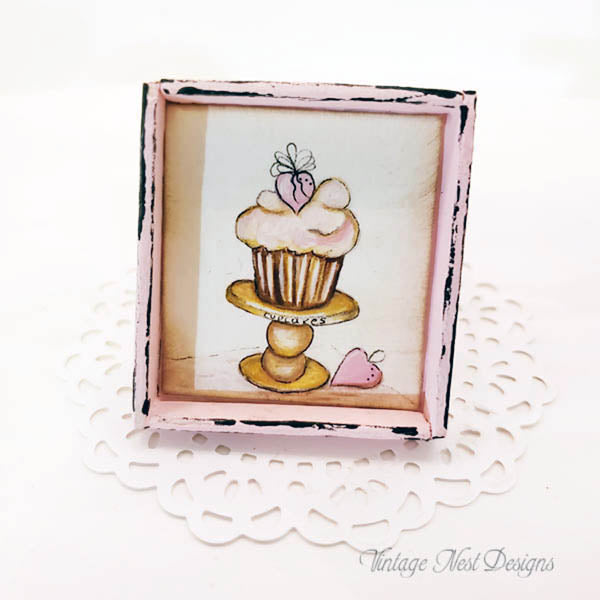 A square framed art print featuring a whimsical cupcake on a pedestal with a small strawberry, displayed on a white paper doily. The frame has a distressed pink and black border.