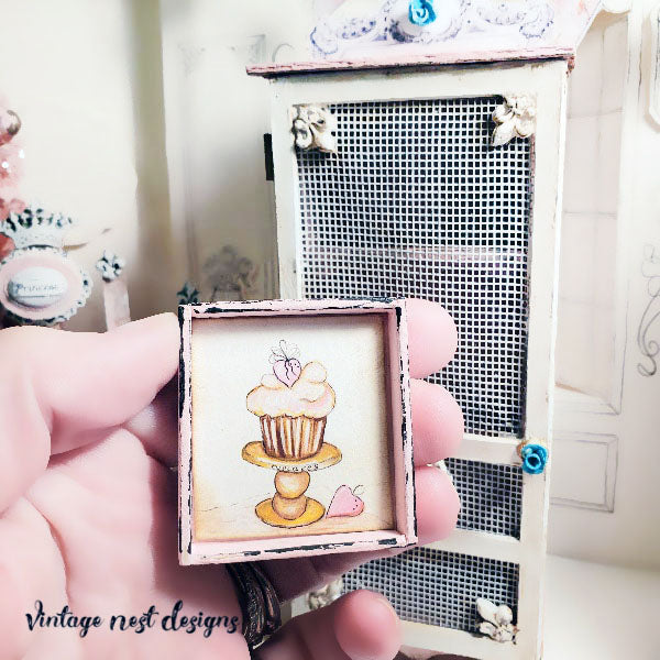 A hand holds a small square decorative frame featuring a cupcake illustration, with a miniature cabinet in the background. The watermark 'Vintage nest designs' appears in the lower left corner.