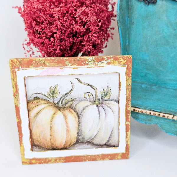 Small painting of pumpkins in a rustic frame with pink flowers and a blue vase in the background.