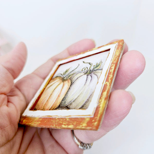 Hand holding a small framed painting of pumpkins against a white background