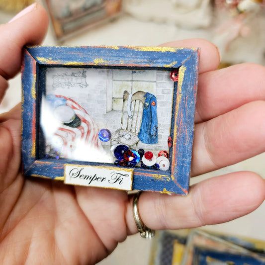 A hand holding a small rectangular decorative shadow box. The frame is distressed blue with gold paint details and a small white label at the bottom that reads 'Semper Fi'.