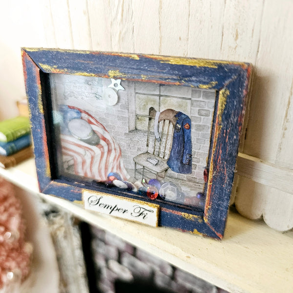 A small rectangular decorative frame with a distressed blue and gold border, containing a miniature scene of a chair, coat, and American flag, displayed on a shelf with a sign reading 'Semper Fi'.