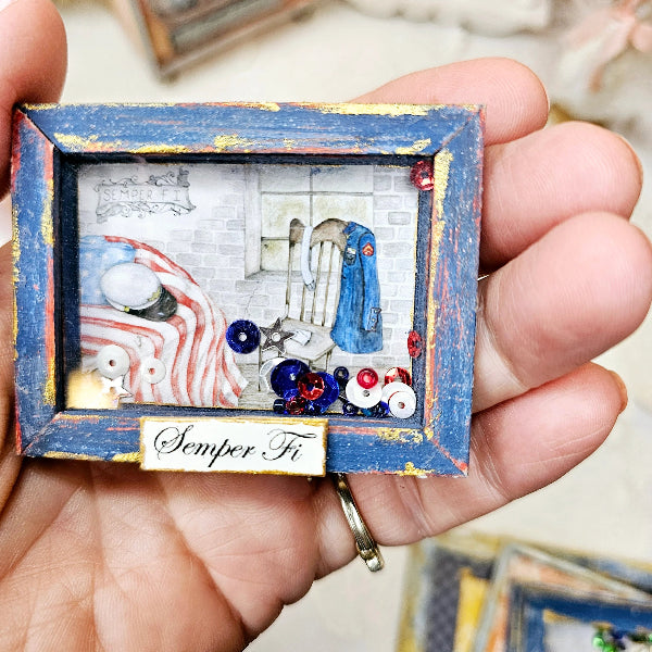 A hand holding a small decorative shadow box plaque with a distressed blue frame, patriotic red, white, and blue embellishments, and a white label reading 'Semper Fi'.