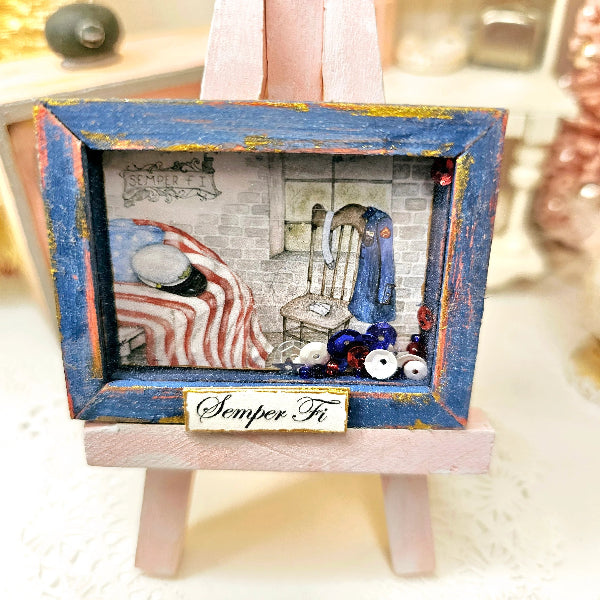 A miniature dollhouse-scale framed shadow box canvas on a pink display easel. The artwork features a blue distressed frame with gold paint splatters, containing a detailed miniature scene of a bedroom with a striped blanket, a blue coat, and a brick wall, with a 'Semper Fi' plaque at the bottom.
