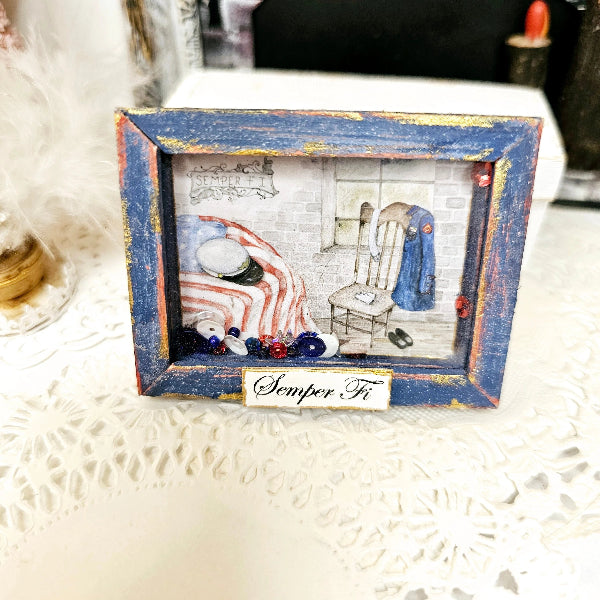 A square decorative shadow box plaque with a distressed blue and gold frame, featuring a miniature interior scene with a striped bed, chair, and blue coat, plus a small label reading 'Semper Fi' on a white lace surface.