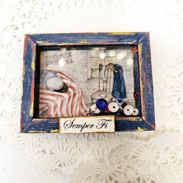 A square framed shadow box with a distressed blue and gold frame, containing a patriotic-themed collage with red and white stripes, a blue chair, and spools of thread. A small label at the bottom reads 'Semper Fi'.