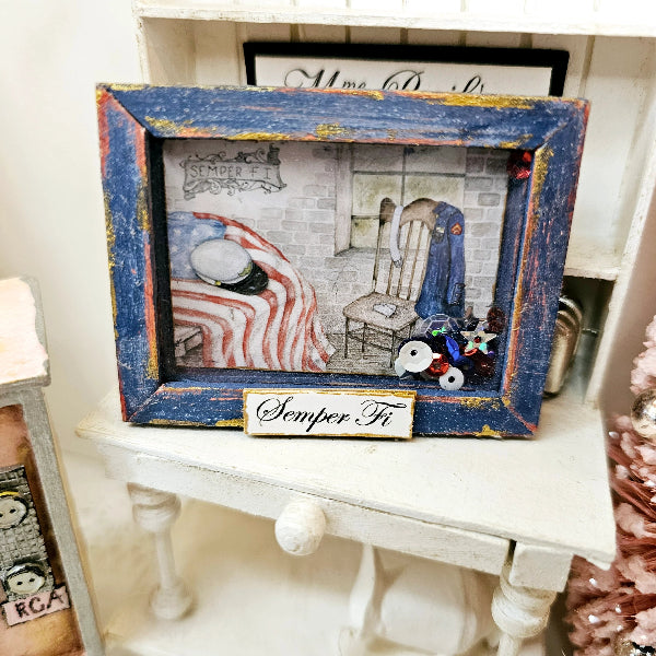A distressed blue wooden picture frame with gold and red paint chipping, containing a patriotic-themed miniature scene with an American flag, a chair, and a small 'Semper Fi' plaque, displayed on a white miniature table.