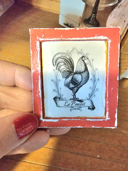 Small framed artwork of a rooster with 'Le Poulet' text held by a person.