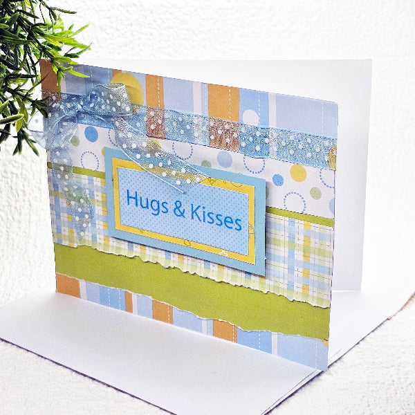 A handmade greeting card with a blue and yellow patterned design, featuring the text 'Hugs & Kisses' on a blue label, set against a white background with a green plant leaf in the corner.