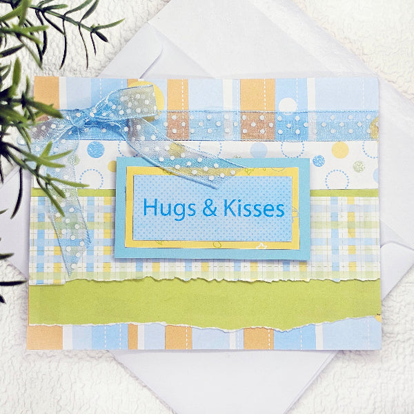 A handmade greeting card with layered pastel patterned paper in blue, yellow, and white, featuring a blue ribbon bow and a central label that reads 'Hugs & Kisses' in blue text. The card is displayed on top of a white envelope with a small green plant branch in the corner.