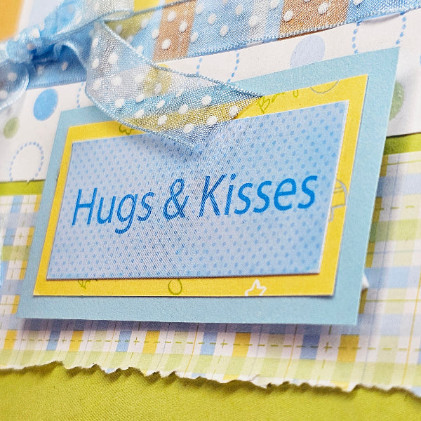 A close-up of a handmade paper tag with the words 'Hugs & Kisses' in blue script, layered over patterned paper in pastel blue, yellow, and white, with a blue polka-dot ribbon tied at the top.