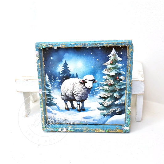 A square decorative framed artwork showing a snowy winter scene with a white sheep standing in front of snow-covered pine trees under a blue night sky. The frame is distressed blue with gold-toned edges, and the piece is displayed against a white background.