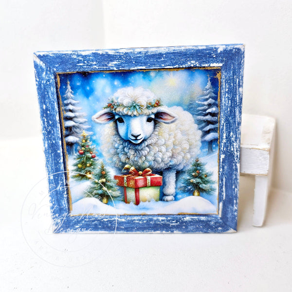 A square framed art print featuring a fluffy white sheep in a snowy winter scene with evergreen trees and a red gift box, set in a distressed blue wooden frame.